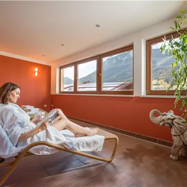 A woman relaxes in a deck chair and reads a book in a cozy room. Large windows offer a view of the mountains in the background.