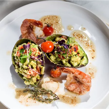 Ein elegantes Gericht mit gefüllten Avocados, Garnelen und einer Kirschtomate. Das Essen ist kreativ angerichtet und mit Kräutern verziert.