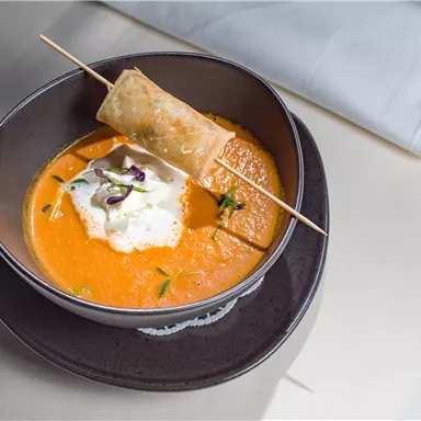 Eine köstliche Karottensuppe mit einem Stück frittiertem Teig und einer Sahnehaube. Serviert in einer eleganten Schüssel auf einem dunklen Teller.
