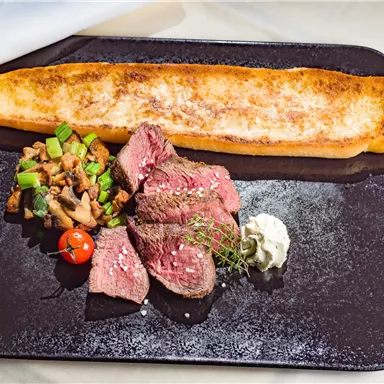 Ein Gourmetgericht mit zartem Rindfleisch, gebratenem Gemüse und einem großen Stück Brot. Das Essen wird von frischen Kräutern und einer Sauce begleitet.