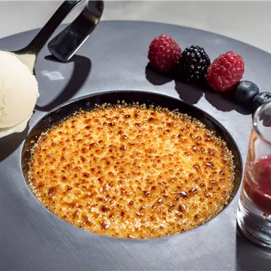 A delicious Crème Brûlée on a stylish plate, served with a scoop of vanilla ice cream and fresh berries. Next to it, a small glass with fruity sauce.