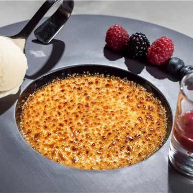 Eine köstliche Crème Brûlée auf einem stilvollen Teller, serviert mit einer Kugel Vanilleeis und frischen Beeren. Daneben ein kleines Glas mit fruchtiger Soße.