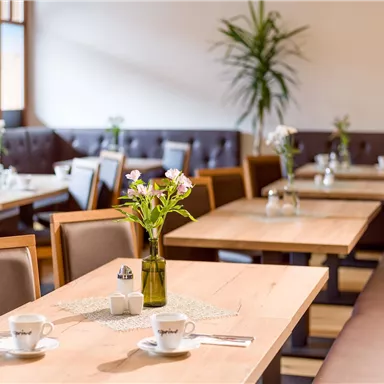 Ein gemütliches Restaurant mit Holztischen und Pflanzen. Die Tische sind für Gäste gedeckt und mit Blumen dekoriert.