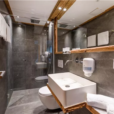 A modern bathroom with elegant gray tiles and wooden details. It features a shower, a sink, and neatly arranged towels.