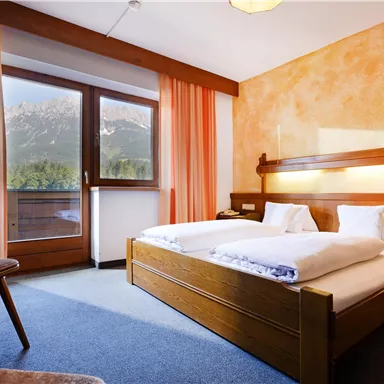 A cozy bedroom with two beds and a wooden desk. Large windows with curtains provide plenty of daylight and a view outside.