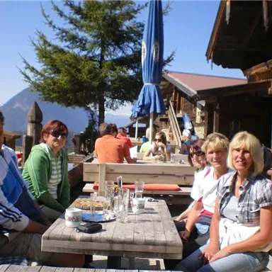 Eine Gruppe von fünf Personen sitzt an einem Holztisch in einer Berghütte. Im Hintergrund sind weitere Gäste und Bäume zu sehen, die die schöne Berglandschaft umgeben.