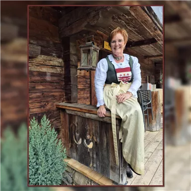 A smiling woman is sitting on a wooden rock in front of a rustic cabin. She is wearing a traditional outfit and has short, reddish-brown hair.