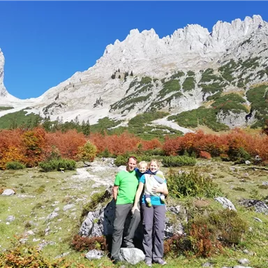 Eine Familie posiert vor einer beeindruckenden Bergkulisse mit buntem Herbstlaub. Der Himmel ist klar und blau, was die natürliche Schönheit der Landschaft hervorhebt.