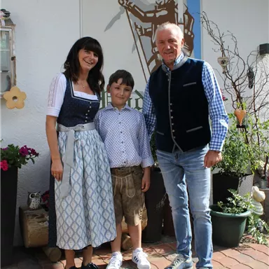 Eine Familie steht fröhlich vor einem Haus mit einer traditionellen Dekoration. Alle tragen traditionelle Kleidung und lächeln in die Kamera.