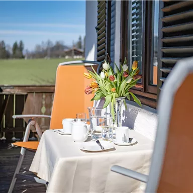 A beautiful balcony with a table set for breakfast. There are flowers, dishes, and glasses on the table.