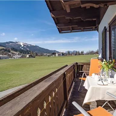 A balcony with a view of a green meadow and the mountains in the background. On the table are flowers and the chairs are orange.