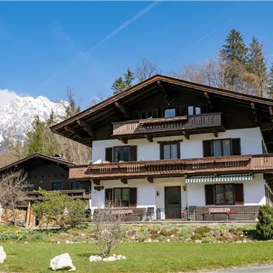 A charming house in alpine style, surrounded by green meadows and mountains in the background. The sky is blue and clear.