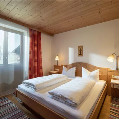 A cozy room with a double bed and two pillows. The room has large windows with a view outside and a wooden ceiling.