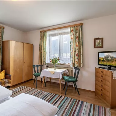 A cozy bedroom with two beds and a small seating area. The room is bright and cheerfully furnished, with a window and a television.