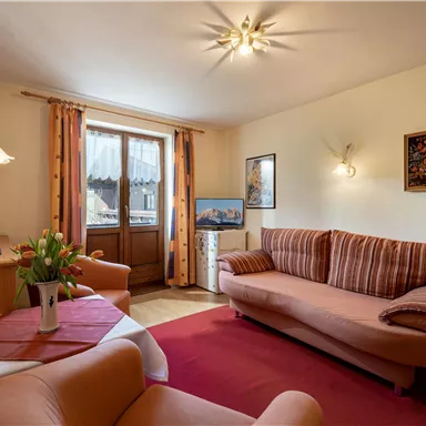 A cozy living room with a red carpet and comfortable sofas. There are windows with curtains and a TV on a small table.