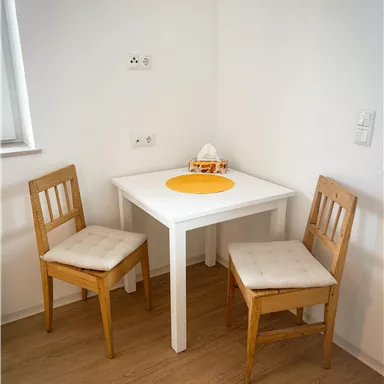 A small dining area with a white table and two wooden chairs. An orange placemat is on the table.