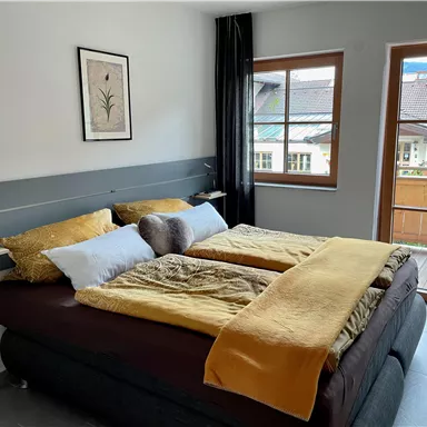 Ein gemütliches Schlafzimmer mit einem großen Bett und warmen Farbtönen. Es gibt ein Fenster mit Blick auf einen Balkon und die umliegenden Gebäude.