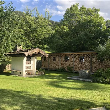 A beautiful garden with a covered seating area and a wood stack wall. In the background, trees and a rural setting can be seen.