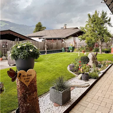 A well-kept garden with green grass, colorful flowers, and various plants. In the background, wooden houses and a cloudy sky can be seen.
