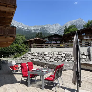 Ein malerischer Außenbereich mit einem Esstisch und roten Stühlen. Im Hintergrund sind majestätische Berge und grüne Bäume zu sehen.