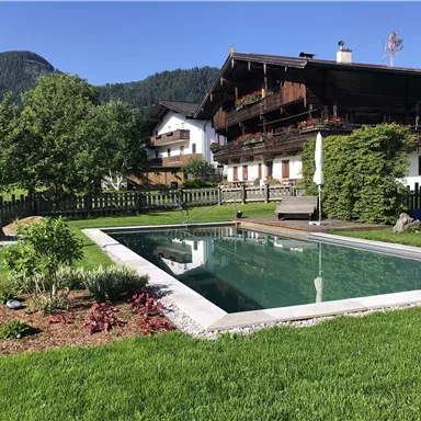 A well-maintained garden with a pool and a wooden terrace. Traditional houses and green mountains can be seen in the background.