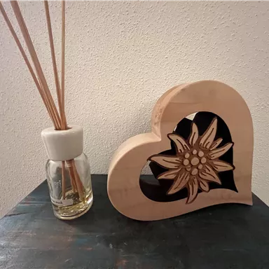 Ein Holzherz mit einer Blume im Inneren steht neben einem Duftbehälter mit langen Stäbchen. Die Szene vermittelt eine gemütliche und dekorative Atmosphäre.