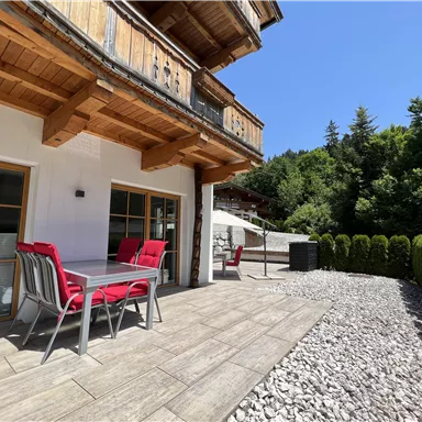 Eine Terrasse mit einem Tisch und roten Stühlen. Im Hintergrund sind Bäume und eine sonnige Himmelstruktur zu sehen.