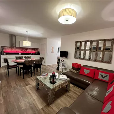 Ein modernes Wohnzimmer mit einem gemütlichen Sofa und einem Esstisch. Die Wände sind hell und es gibt dekorative Elemente mit roten Akzenten.