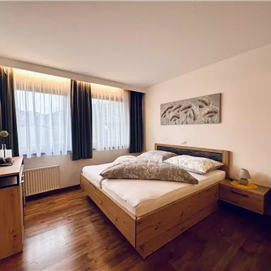 A modern bedroom with a double bed and wood paneling. Large windows let in plenty of daylight.