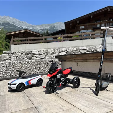 Ein Spielzeugauto und ein Kinderfahrzeug stehen auf einer Terrasse bei schönem Wetter. Im Hintergrund sind Berge und ein modernes Haus zu sehen.