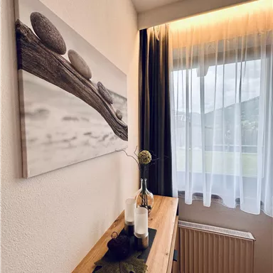 A cozy room with a wooden table and decoration made of stones and a vase. The window is adorned with a white curtain and lets in soft light.