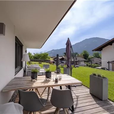 A cozy outdoor area with a wooden table and modern chairs. In the background, green meadows and mountains can be seen.