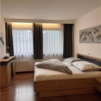 A cozy bedroom with a large bed and wooden flooring. Large windows with curtains let in plenty of light.