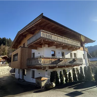 Ein modernes Haus im alpinen Stil mit Holzverkleidung und mehreren Balkonen. Die Umgebung ist von grünen Bäumen und Bergen umgeben.
