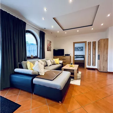A modern living room with a black sofa, colorful cushions, and a large window. The floor is tiled with terracotta-colored tiles, and there is a small wooden table.