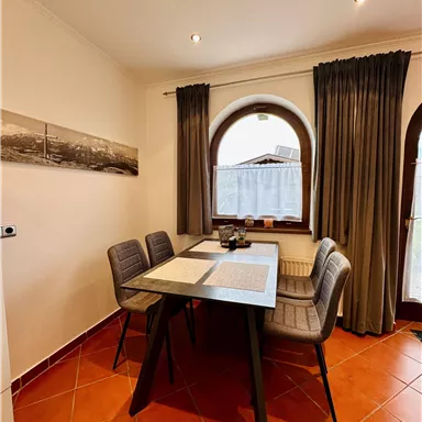 A modern dining room with a table and four chairs. Large windows with curtains provide plenty of light.