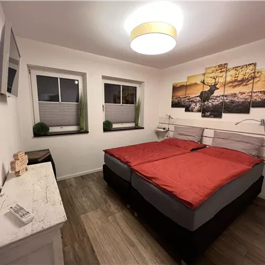 Ein modernes Schlafzimmer mit einem Doppelbett und roten Bettdecken. Die Wände sind mit dekorativen Bildern geschmückt und es gibt Fenster, die Tageslicht hereinlassen.