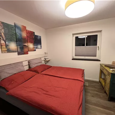 Ein gemütliches Schlafzimmer mit einem Doppelbett und roten Bettdecken. An der Wand hängen farbige Bilder, und das Zimmer ist hell und modern eingerichtet.