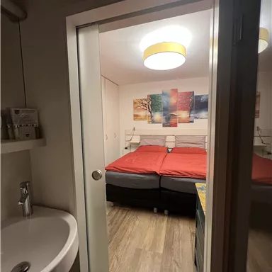 Ein gemütliches Schlafzimmer mit zwei roten Betten und einem modernen Deckenlicht. Im Vordergrund ist ein Waschbecken sichtbar.