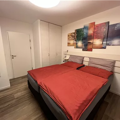 Ein gemütliches Schlafzimmer mit einem großen Doppelbett und roten Bettdecken. An der Wand hängen farbenfrohe Bilder, und es gibt einen kleinen Tisch und einen Kleiderschrank.