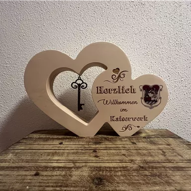 Eine herzförmige Holzdeko mit dem Schriftzug "Herzlich Willkommen im Katzenwerk" und einem Schlüssel. Die Dekoration steht auf einem Holztisch und verleiht einen einladenden Eindruck.