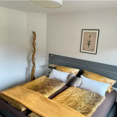 Ein gemütliches Schlafzimmer mit einem großen Doppelbett und gelben Bettdecken. An der Wand hängt ein dekoratives Bild, und eine Holzskulptur steht im Raum.