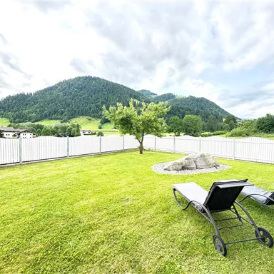 A green meadow with two loungers and a view of gentle hills. In the background, mountains and a blue sky can be seen.