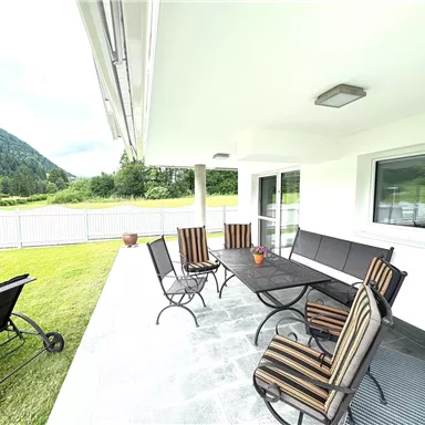 A cozy outdoor area with a table and several chairs. In the background, green meadows and mountains can be seen.
