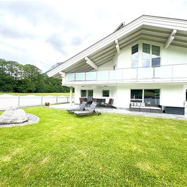 A modern house with large windows and a spacious terrace. The garden is well-maintained and offers a green lawn area.