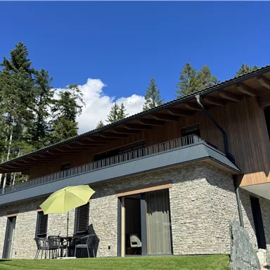 A modern house with a stone and wooden facade, surrounded by trees. On the terrace, there are tables and chairs as well as a yellow sun umbrella.