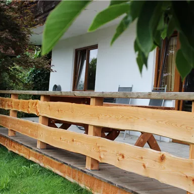 A wooden terrace with a beautiful railing surrounds the house. In the background, windows and green plants can be seen.