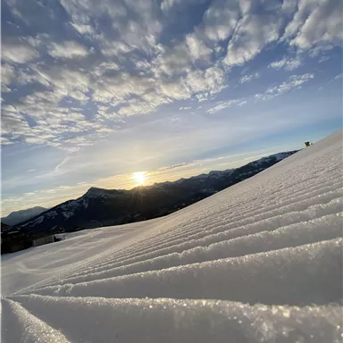 Eine verschneite Landschaft mit sanften Hügeln, die von Sonnenlicht beleuchtet wird. Der Himmel ist klar und weist einige Wolken auf.