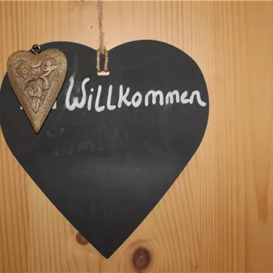A heart-shaped sign made of black material with the inscription "Welcome". It hangs on a wooden surface and has a small wooden tag.