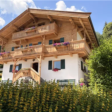 A traditional wooden house with flower balconies and a green garden. The sky is clear and a few clouds are visible.